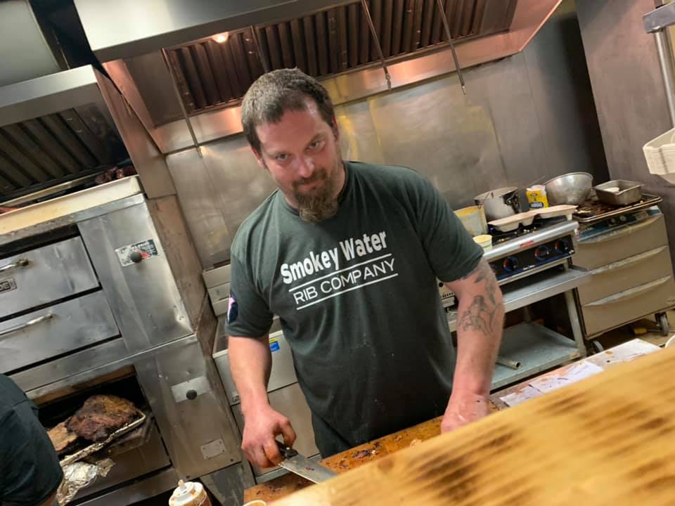 Pitmaster in a Smokey Water Rib Company tee slicing brisket in the kitchen.
